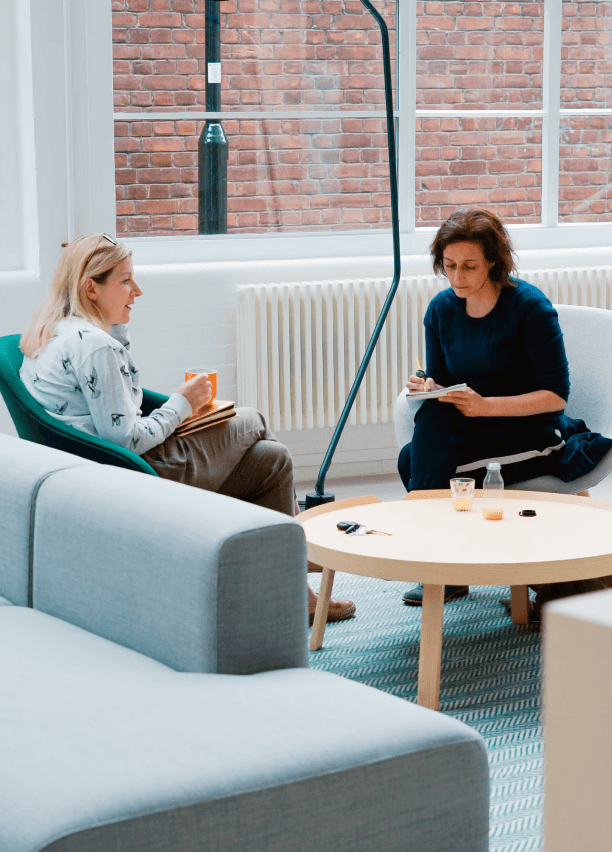 Two women are sitting and talking in a modern office lounge with a round wooden table and gray couches, while one of them writes on a notepad.
