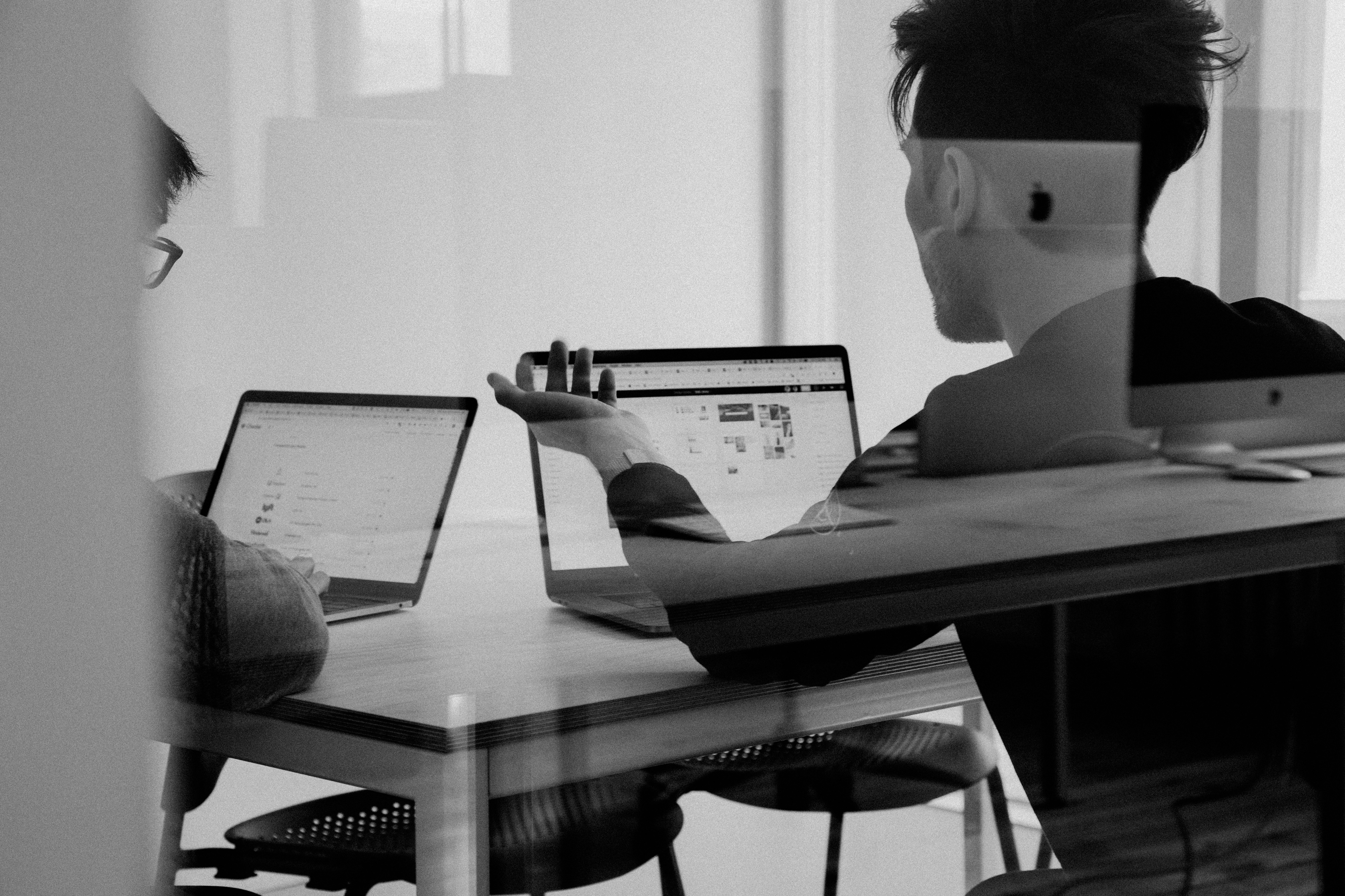 Two people sit at a wooden table, engaged in a discussion with laptops open, in a modern office setting illuminated by natural light.