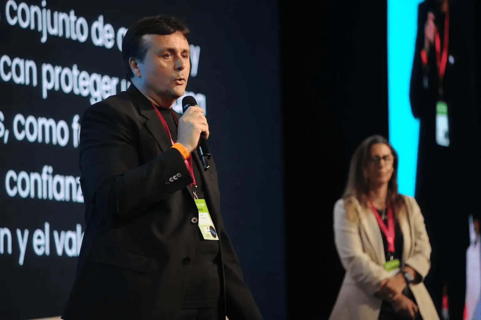 A man in a black suit is speaking into a microphone on stage at a conference, while a woman in a beige jacket stands in the background; a large screen displaying text forms part of the backdrop.