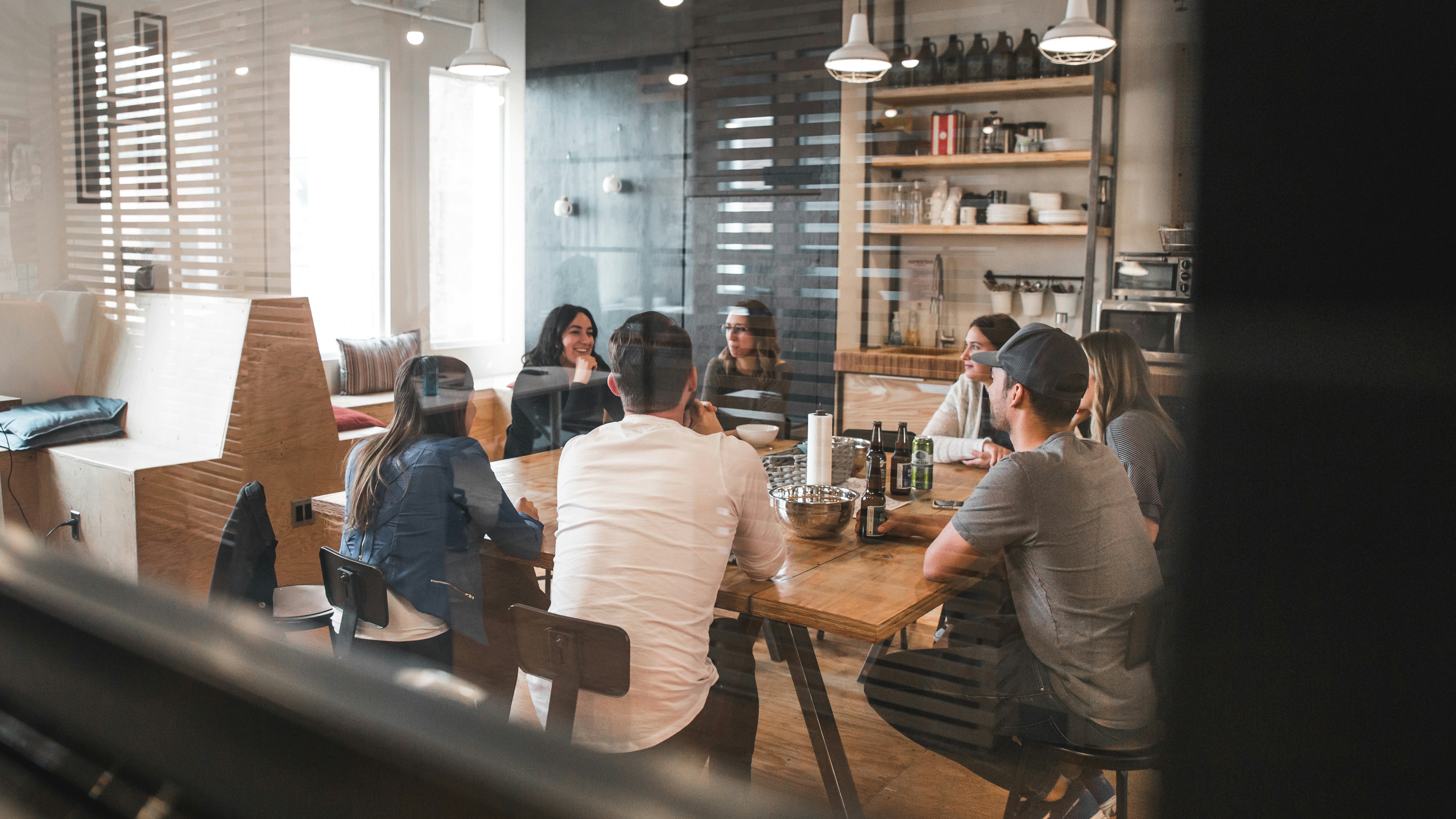 A group of people are seated at a wooden table in a modern, industrial-style meeting room with shelves and pendant lights, engaged in a discussion.